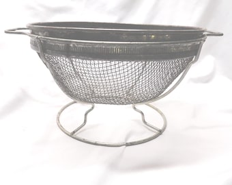 May include: A vintage metal mesh strainer with a circular base and two handles. The strainer has a fine mesh and a dark rim. The overall color is silver and gray, suggesting it is made of stainless steel or a similar metal.