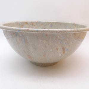 May include: A round mixing bowl with a wide, flat rim and a smooth, glossy finish. The bowl features a speckled, marbled pattern in shades of beige, tan, and light brown, with small, scattered flecks of blue and white throughout the surface.