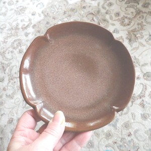 May include: A brown ceramic saucer with a scalloped edge. The saucer has a speckled glaze and a smooth, slightly textured surface.