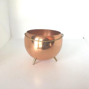 May include: A copper planter with a brass rim and three brass legs. The planter is round and has a shiny finish.