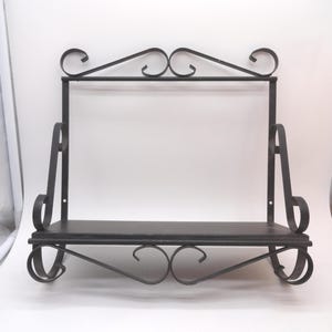 May include: A wall-mounted shelf featuring a dark metal frame with decorative scrollwork accents. The unit includes a single flat rectangular shelf surface supported by curved metal brackets and a top horizontal bar with ornate metal curls.