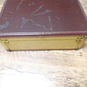 May include: A rectangular suitcase featuring a dark brown top lid with visible surface scuffs and a cream-colored base. The case includes gold-toned metal hinges and corner reinforcements along the edges, showcasing a two-tone design with visible stitching.