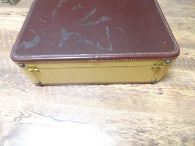 May include: A rectangular suitcase featuring a dark brown top lid with visible surface scuffs and a cream-colored base. The case includes gold-toned metal hinges and corner reinforcements along the edges, showcasing a two-tone design with visible stitching.