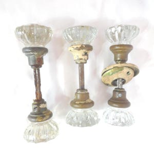 May include: Three antique glass doorknobs with aged metal hardware. The clear glass knobs have a ribbed design. The metal components show signs of wear, with a mix of brass and rusted finishes. A vintage hardware set.