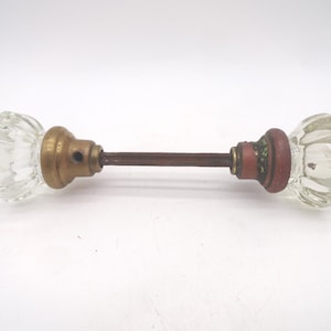 May include: A door knob set featuring two clear, fluted glass knobs connected by a central metal spindle. One knob is attached to a brass-toned circular base with a small hole, while the other is attached to a dark, weathered red metal base.