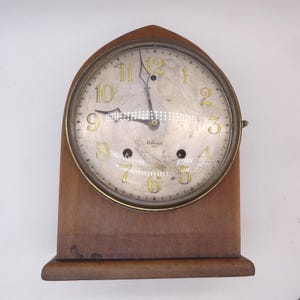 May include: A wooden mantel clock with a pointed arch top and a circular face. The clock features a silver-toned dial with gold-colored numerals, black hands, and the text Gilbert printed on the face. Two winding holes are visible on the dial.
