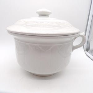 May include: A white ceramic lidded serving dish with a handle. The dish has a round shape with a decorative pattern and a matching lid with a knob. The handle is curved. The dish is likely for serving food.