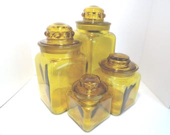 May include: Four amber-colored glass canisters with matching lids. The canisters are square-shaped and vary in size, creating a decorative set. The lids have a decorative design. The glass has a slight tint of yellow.
