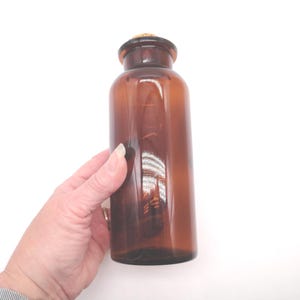 May include: A cylindrical amber glass bottle with a narrow neck and a rounded shoulder. The bottle features a smooth, translucent brown finish and is topped with a small, light-colored cork stopper that fits securely into the circular opening.