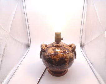 Op de afbeelding: Een bruine keramische tafellamp met een lamphouder en een bruin elektrisch snoer. De lamp heeft een ronde behuizing met twee decoratieve handvatten en een bruine voet. De lamp is verlicht en geeft een warme gloed.