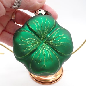 May include: A green glass ornament shaped like a four-leaf clover, with glitter accents. The ornament is held by a hand and sits on a copper-colored base. The clover design is a symbol of good luck.