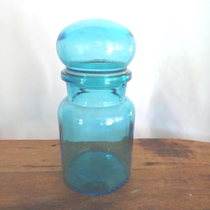 May include: A turquoise glass jar with a rounded lid. The jar has a cylindrical body and a slightly flared base. The lid is a rounded shape. The jar is sitting on a wooden surface.