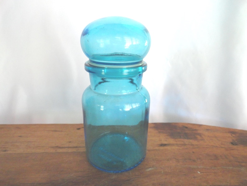 May include: A turquoise glass jar with a rounded lid. The jar has a cylindrical body and a slightly flared base. The lid is a rounded shape. The jar is sitting on a wooden surface.