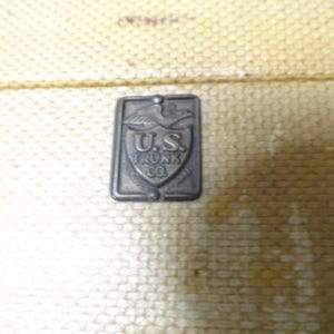 May include: A rectangular metal emblem attached to a textured, light tan surface. The emblem features an eagle at the top, a shield shape in the center, and the embossed text U.S. TRUNK CO. The metal has a dark, weathered, and aged appearance.