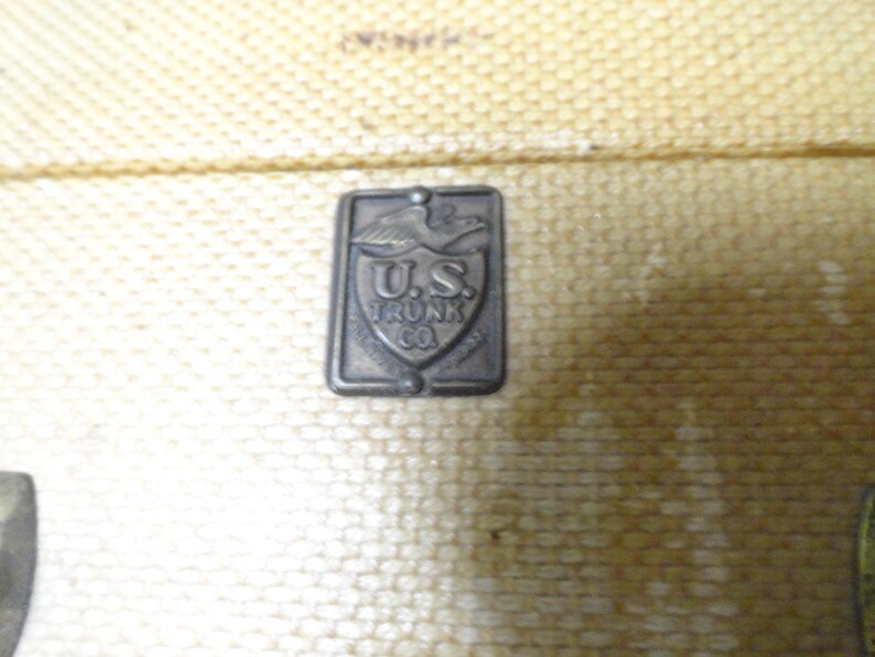 May include: A rectangular metal emblem attached to a textured, light tan surface. The emblem features an eagle at the top, a shield shape in the center, and the embossed text U.S. TRUNK CO. The metal has a dark, weathered, and aged appearance.