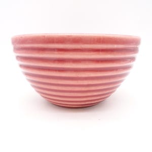 May include: A small, round, pink ceramic bowl with horizontal ribbed detailing. The bowl has a smooth rim and a slightly darker shade of pink at the base. The bowl is set against a white background.