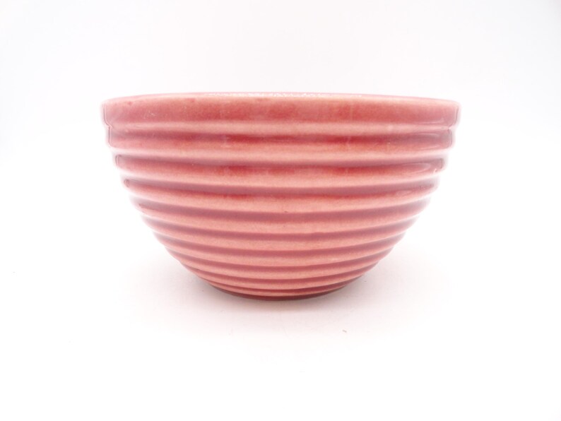 May include: A small, round, pink ceramic bowl with horizontal ribbed detailing. The bowl has a smooth rim and a slightly darker shade of pink at the base. The bowl is set against a white background.