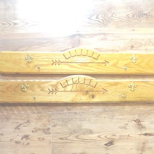 May include: Two wooden wall-mounted coat racks. Each rack is light brown with a carved arrow and sunburst design. Gold-colored fleur-de-lis and small hooks are also attached. The racks are displayed on a wooden surface.