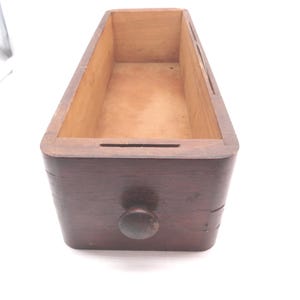 May include: A rectangular wooden drawer with a dark brown finish and a single centered round knob on the front panel. The interior of the drawer is a lighter natural wood tone, and the top edges feature narrow horizontal slots on each side.