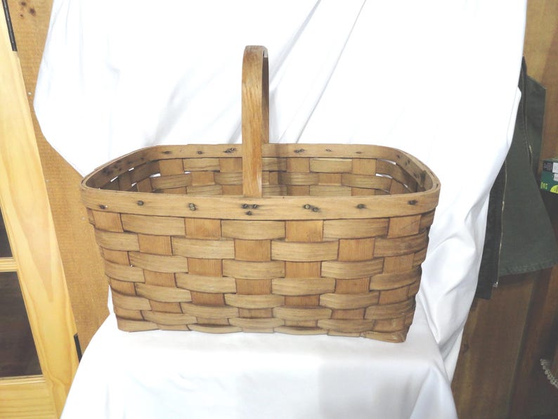 May include: A rectangular wooden basket with a woven design and a single wooden handle. The basket is made of natural wood and has a rustic appearance.