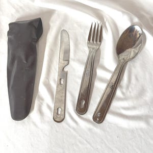 May include: A stainless steel camping utensil set includes a knife with a bottle opener, a fork, and a spoon. The set comes with a black nylon carrying case. The utensils are designed for outdoor use.