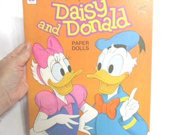 May include: Vintage Disney Daisy and Donald Paper Dolls book with an orange cover. The cover features illustrations of Daisy Duck and Donald Duck. The title "Daisy and Donald" is in purple and yellow lettering. The words "Paper Dolls" are also on the cover.