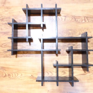 May include: A set of four black wooden wall shelves in various geometric shapes. The shelves are arranged on a light brown wooden floor. The shelves are designed with a grid-like structure, perfect for displaying items.
