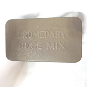 May include: A rectangular, light brown tin with rounded corners. The embossed text on the lid reads "DROMEDARY DIXIE MIX". The tin appears to be made of metal and has a slightly textured surface.