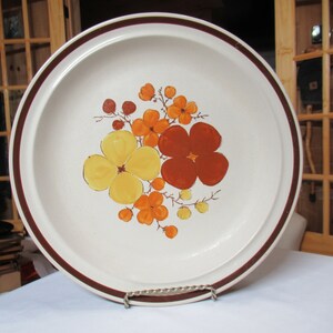 May include: A white ceramic dinner plate with a brown rim and a floral design featuring yellow, orange, and red flowers.