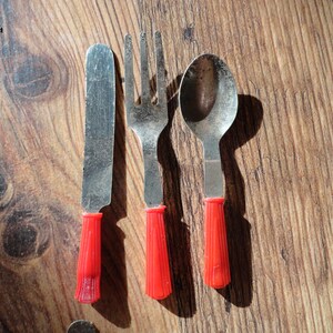 May include: A set of three red-handled silverware pieces: a knife, fork, and spoon. The silverware is made of metal and has a vintage look.