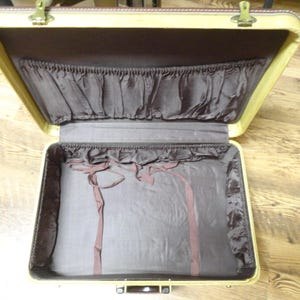 May include: An open vintage suitcase with a cream-colored exterior frame and a dark brown fabric interior lining. The lid features a gathered pocket, while the base includes matching brown fabric side panels and two thin fabric tie-down straps.