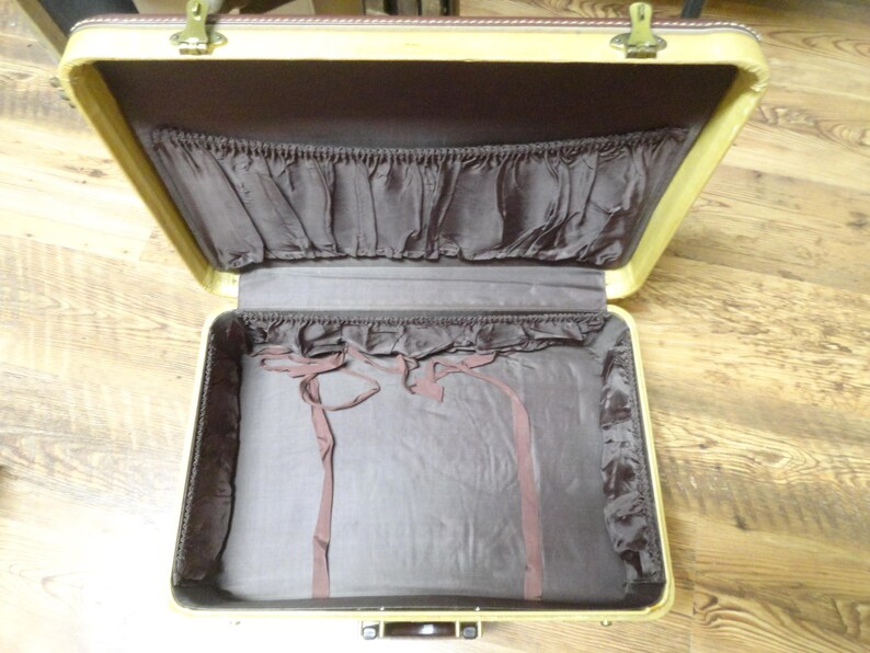 May include: An open vintage suitcase with a cream-colored exterior frame and a dark brown fabric interior lining. The lid features a gathered pocket, while the base includes matching brown fabric side panels and two thin fabric tie-down straps.