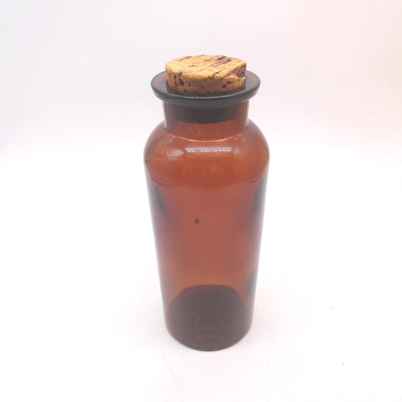 May include: A cylindrical amber glass bottle with a smooth, translucent finish and a wide neck opening. The bottle is sealed with a round, textured natural cork stopper that fits securely into the top rim, providing a simple, functional closure.