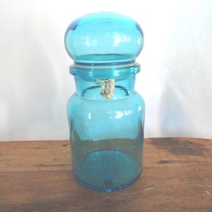 May include: A vintage, turquoise-colored glass apothecary jar with a rounded lid. The jar has a cylindrical body and a wide mouth, with a metal clasp. The jar is sitting on a wooden surface.