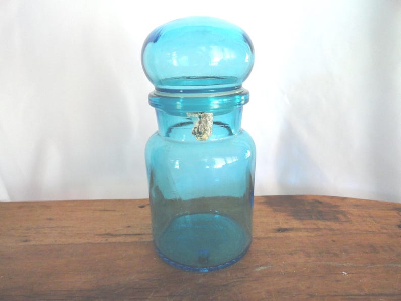 May include: A vintage, turquoise-colored glass apothecary jar with a rounded lid. The jar has a cylindrical body and a wide mouth, with a metal clasp. The jar is sitting on a wooden surface.