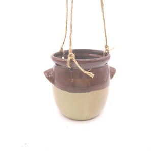 May include: A hanging ceramic planter with a two-tone design. The top portion is a dark brown, transitioning to a light beige at the base. It features small side handles and is suspended by natural twine.