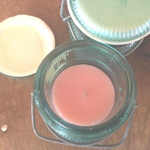 May include: A glass jar candle with a pink candle inside and a metal lid. The jar has a green glass exterior with a decorative pattern.