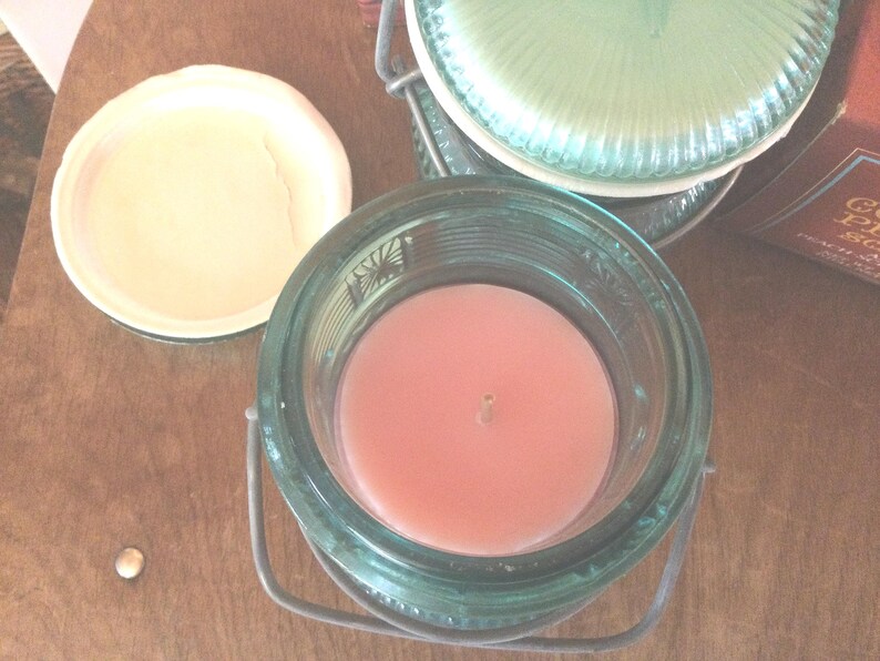 May include: A glass jar candle with a pink candle inside and a metal lid. The jar has a green glass exterior with a decorative pattern.