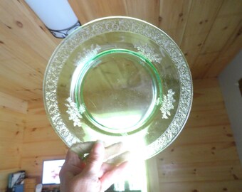 May include: A round, translucent light green glass plate featuring a decorative embossed floral pattern along the wide rim. The center of the plate is smooth and clear, while the outer edge displays a detailed, repeating botanical design.
