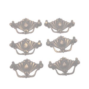 May include: Six antique bronze drawer pulls with a decorative shell design. Each pull features a curved handle and ornate detailing, perfect for adding a vintage touch to furniture. The pulls are made of metal and have a warm, aged finish.