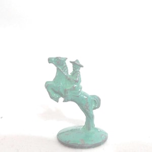 May include: A small, vintage, turquoise-colored toy cowboy riding a rearing horse. The cowboy is wearing a hat and is seated on the horse. The toy is made of metal and has a weathered appearance, suggesting it is an antique.