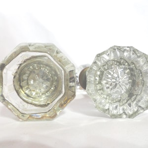 May include: Two clear glass door knobs with a faceted design. One knob is octagonal, and the other is round with a starburst pattern. The knobs are likely vintage and suitable for home decor.