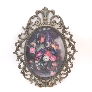 May include: An oval wall frame with an ornate, antique brass-colored metal border featuring intricate scrollwork and leaf motifs. The frame encloses a dark-toned print depicting a vibrant bouquet of multi-colored flowers including pink, red, and white blooms.