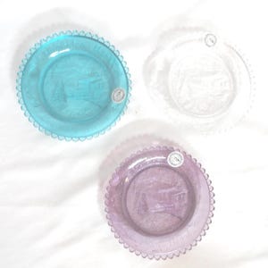 May include: Three decorative glass plates in shades of blue, clear, and purple. Each plate has a scalloped edge and a detailed embossed design of a building and trees. The plates are likely for display.
