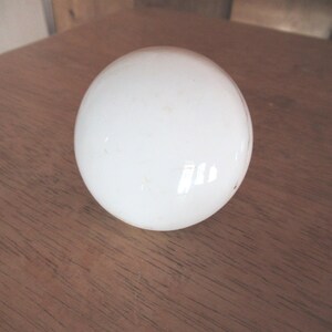 May include: A white, spherical object, possibly a decorative element or part of a larger item, rests on a wooden surface. The sphere has a smooth, reflective surface, with subtle imperfections. The wood grain is visible, adding texture to the scene.