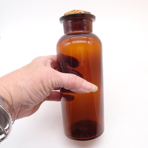 May include: A cylindrical amber glass apothecary bottle with a wide neck and a natural cork stopper. The glass is translucent and brown, featuring a smooth, rounded body and a flat base. The bottle is empty and clear, showing light reflections.
