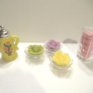 May include: A yellow ceramic teapot with a floral design, three glass dishes with yellow, purple, and green floral candles, and a clear glass jar with pink heart-shaped soaps.