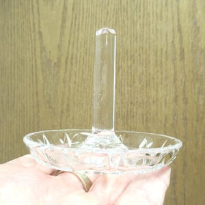 Op de afbeelding: Transparante glazen ringhouder met een ronde, facetgeslepen basis en een lange, rechthoekige paal. De ringhouder wordt in een hand gehouden, met een houten achtergrond.