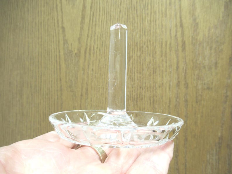 Op de afbeelding: Transparante glazen ringhouder met een ronde, facetgeslepen basis en een lange, rechthoekige paal. De ringhouder wordt in een hand gehouden, met een houten achtergrond.