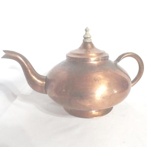 May include: A copper teapot with a rounded body, curved spout, and a small handle. The lid is topped with a decorative finial. The teapot has a polished finish and shows signs of age.
